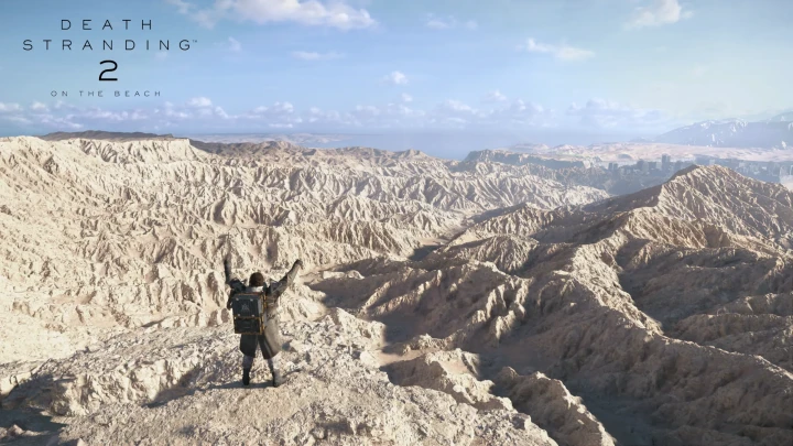 Sam, carrying his cargo backpack, raises his arms on top of pale rock canyons; the sea and a city in the background.