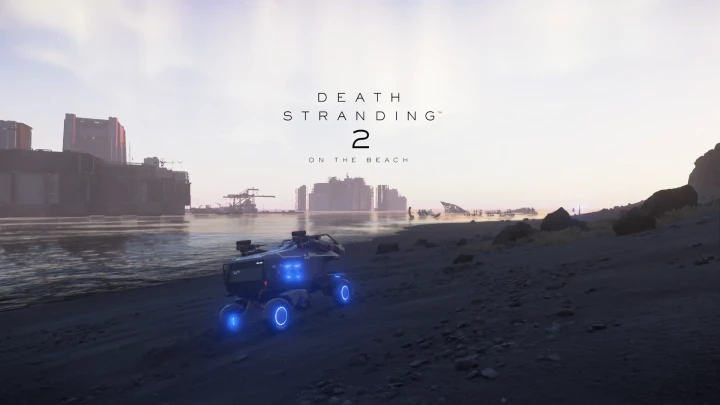 A vehicle with blue lights drives along a black sand beach in front of ruined buildings.